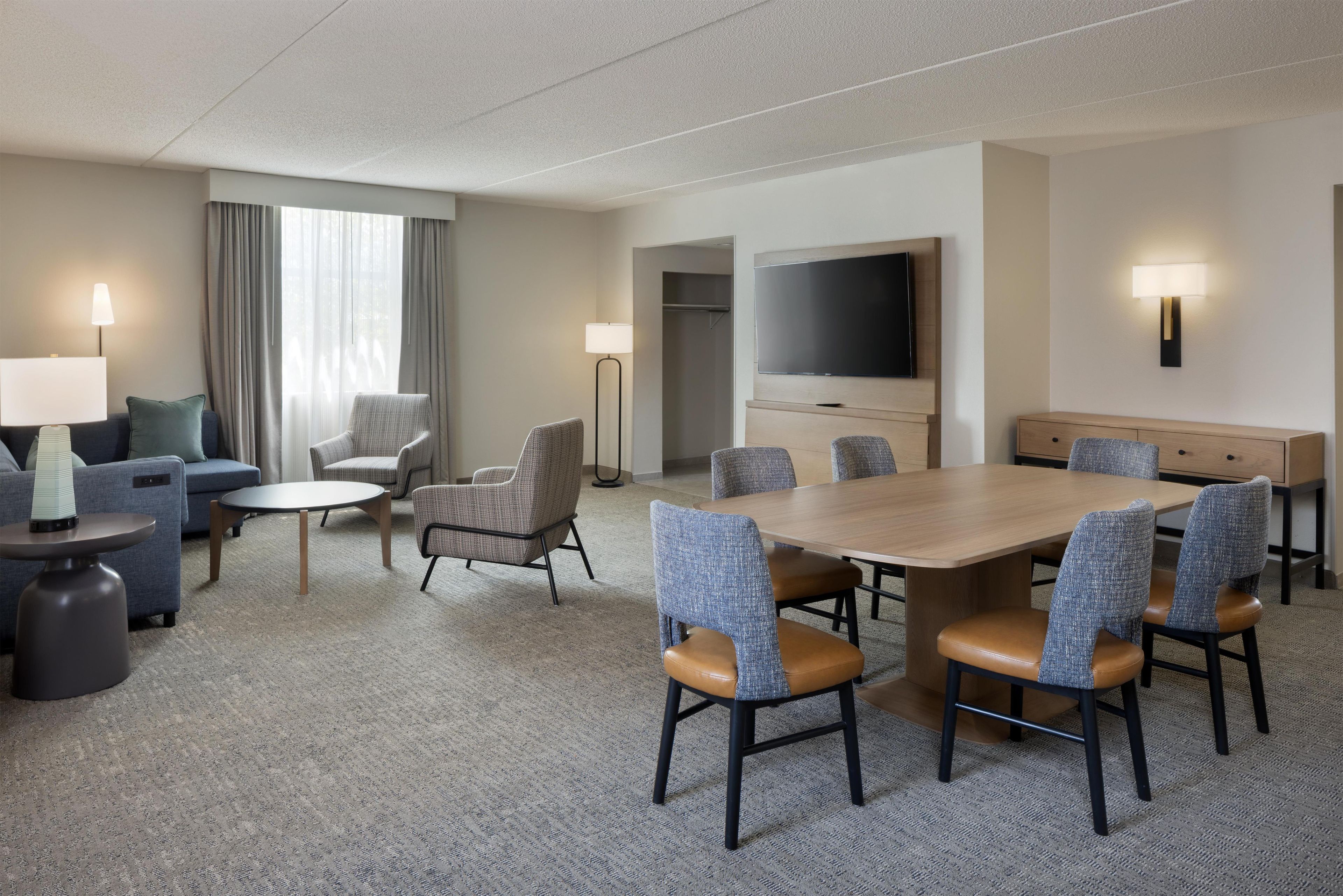 A hotel room with a dining table, chairs, a sofa, a TV, and a lamp.