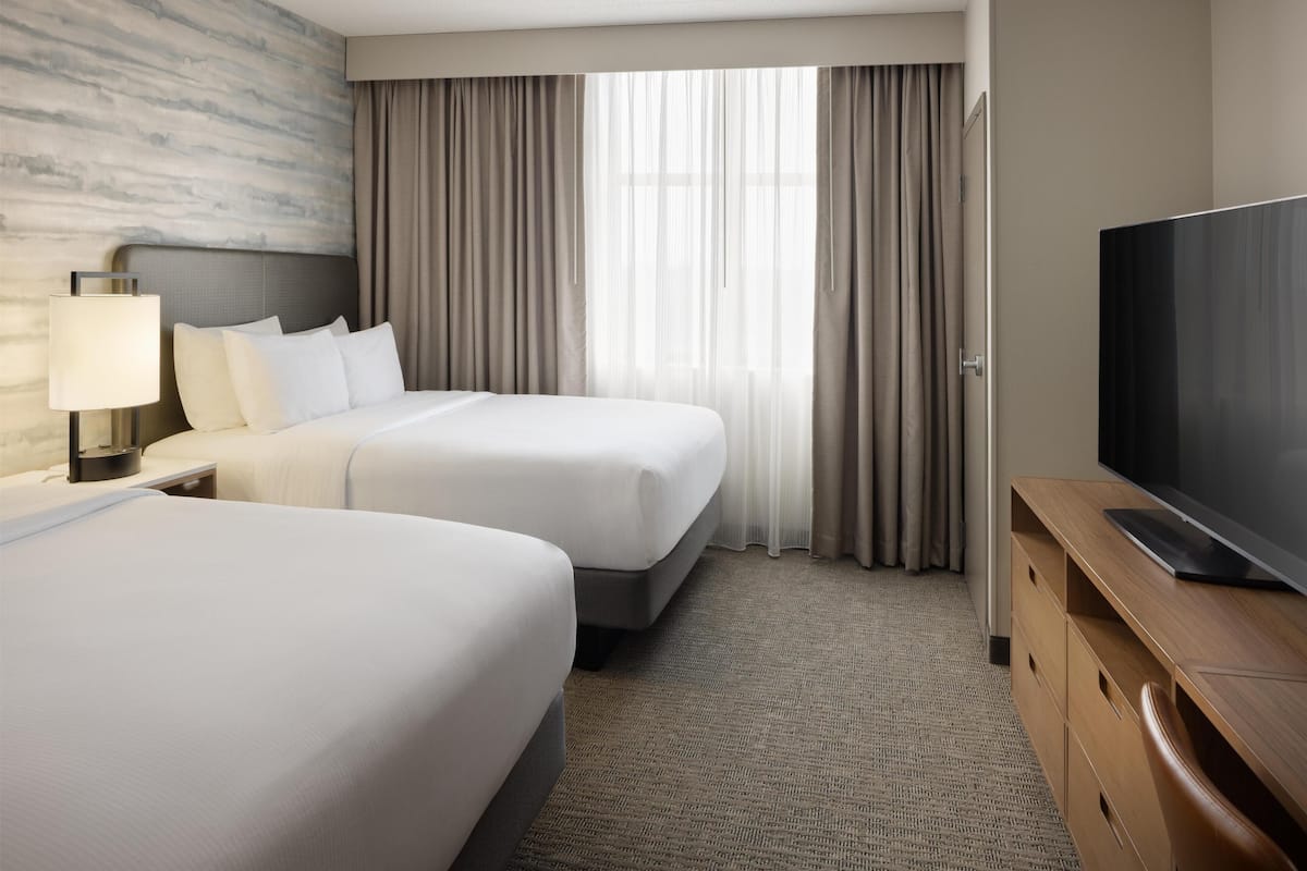 Ein Hotelzimmer mit zwei Betten, einem Fernseher, einer Lampe und einem großen Fenster mit Vorhängen.