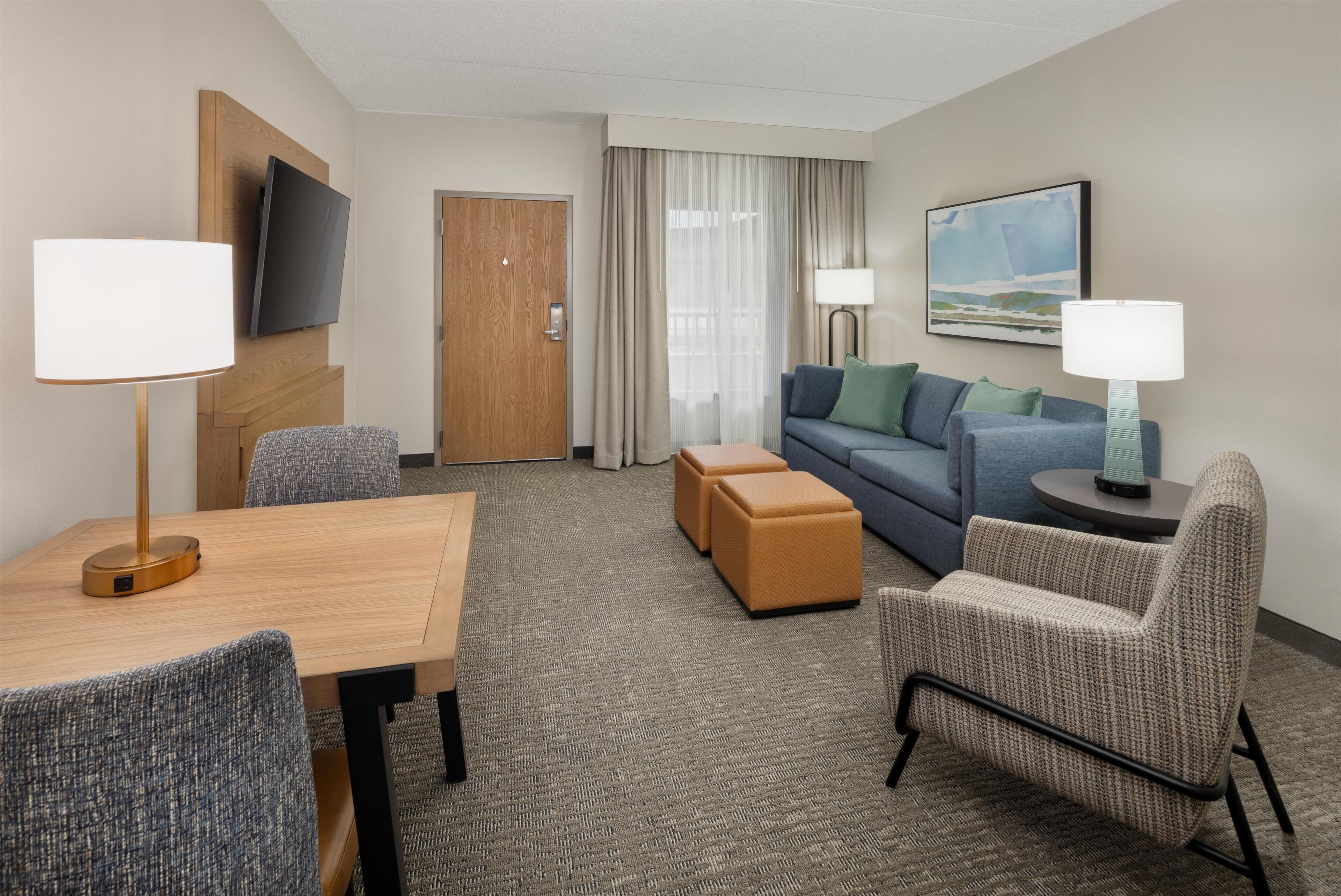 Una habitación de hotel con una mesa de madera, dos sillones, un sofá, un televisor montado en la pared, un cuadro y una puerta.