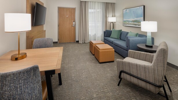 A hotel room with a wooden table, two armchairs, a sofa, a TV mounted on the wall, a painting, and a door. - Embassy Suites by Hilton Lexington/UK Coldstream (Lexington)