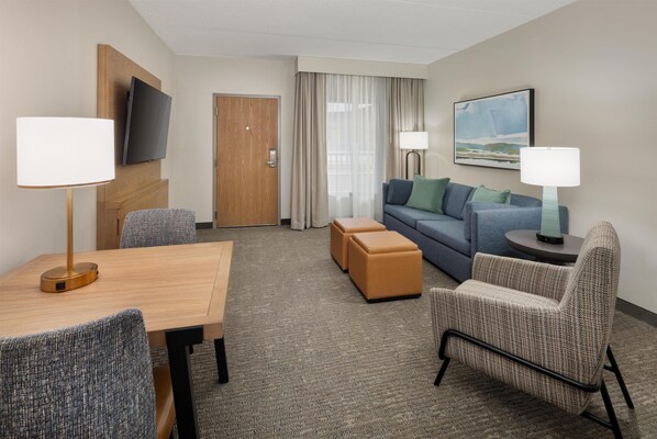 Una habitación de hotel con una mesa de madera, dos sillones, un sofá, un televisor montado en la pared, un cuadro y una puerta.