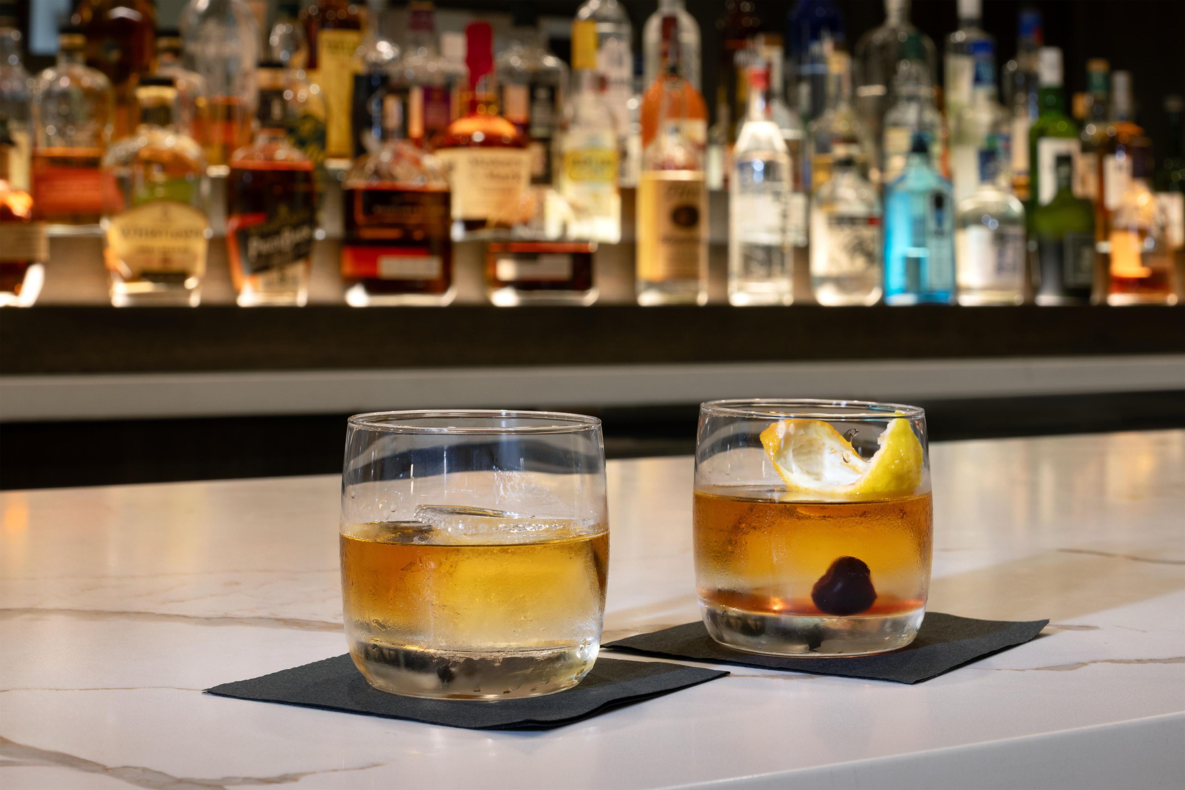 Two glasses of a beverage on a bar counter with a blurred background of various bottles.