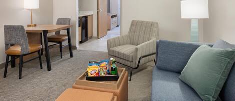 A hotel room with a dining area, a sofa, a chair, and a small table with snacks.