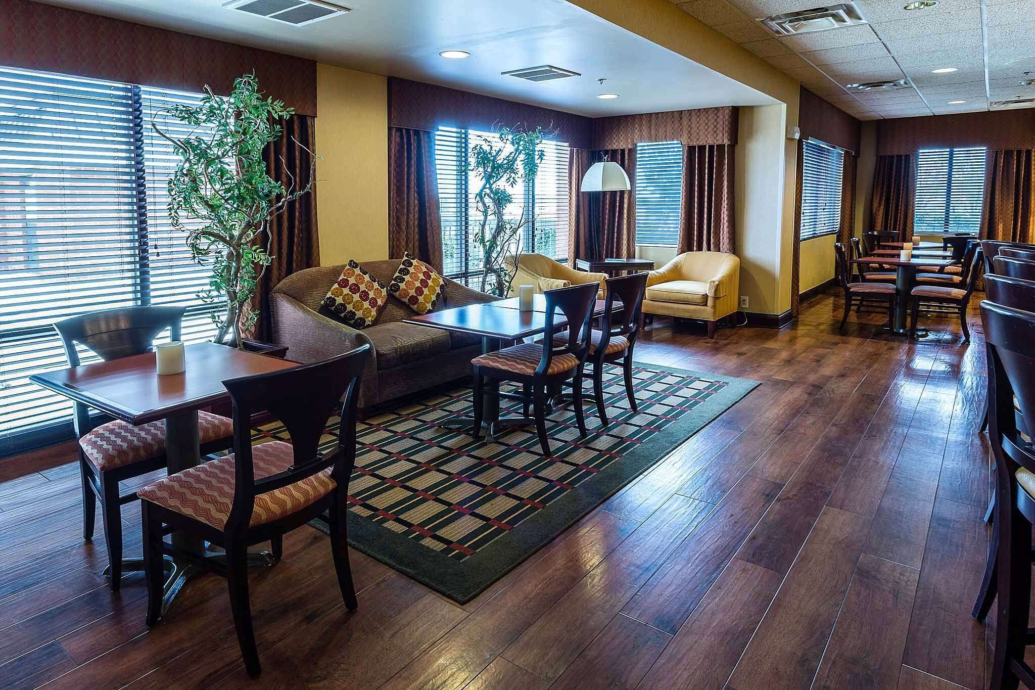 A hotel lobby with a wooden floor, a sofa, a dining table with chairs, and a large potted plant.