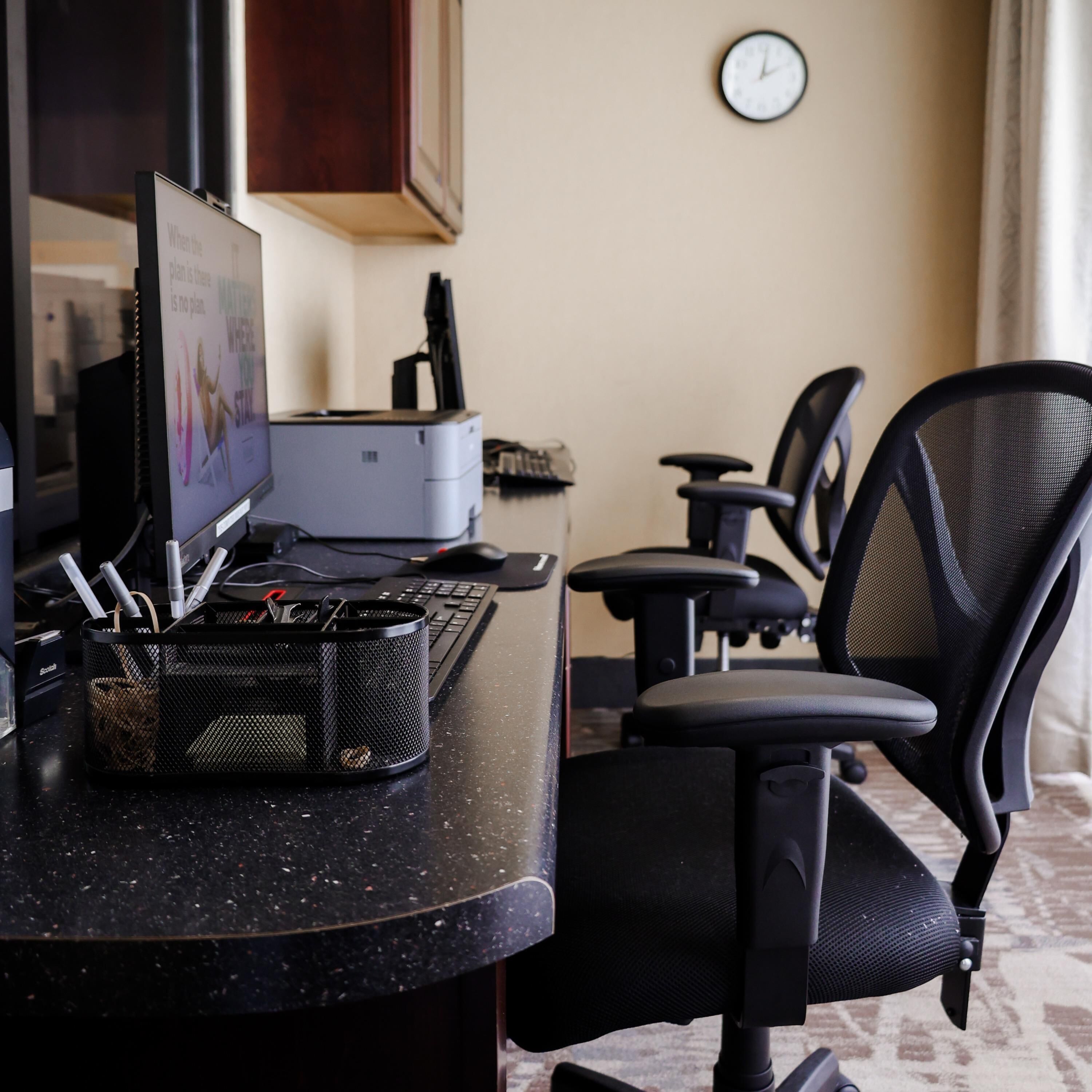 Un espacio de trabajo en un hotel con escritorio, computadora, impresora y sillas de oficina.