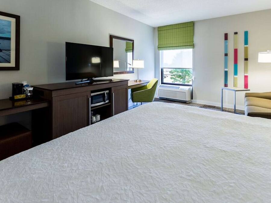 A hotel room with a large bed, a desk with a flat-screen TV, a chair, a window with a view, and a lamp.