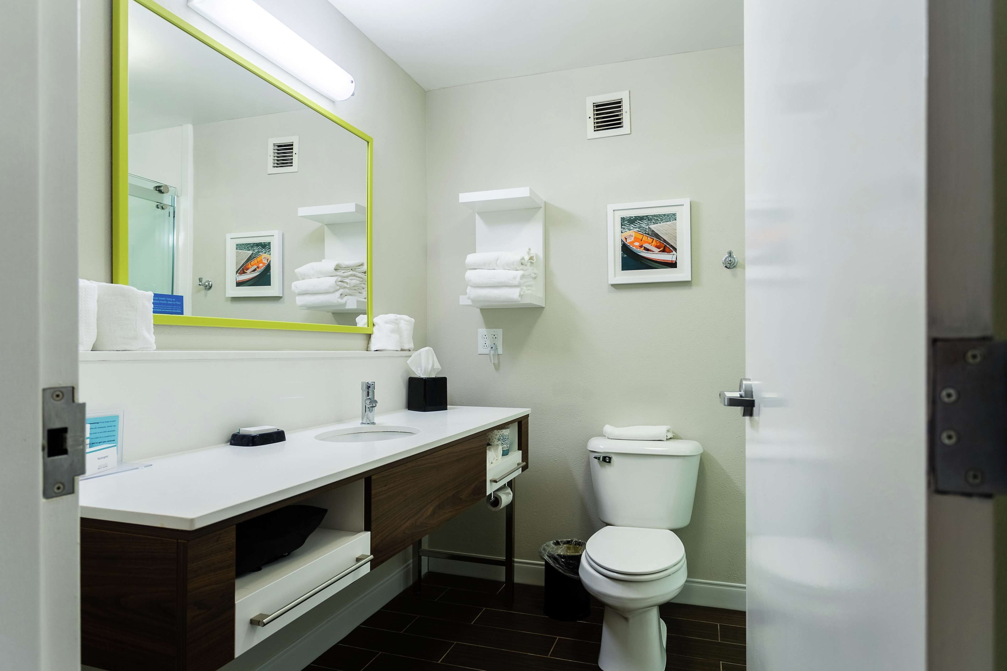 Un baño moderno con lavamanos, espejo y retrete blancos. Hay toallas y un cubo de basura.