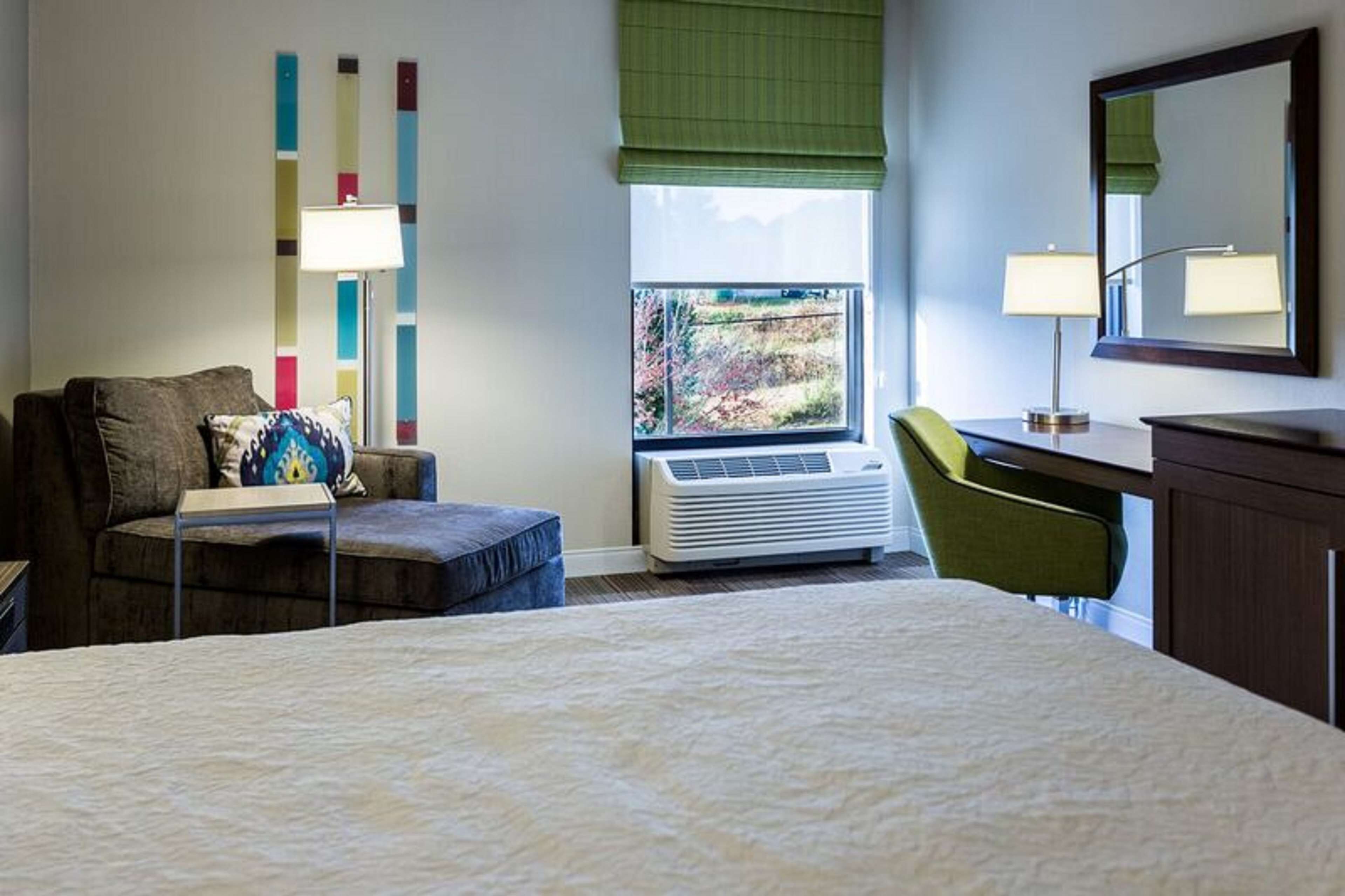 A hotel room with a bed, a desk, a chair, a window with a view, an air conditioner, and a mirror.