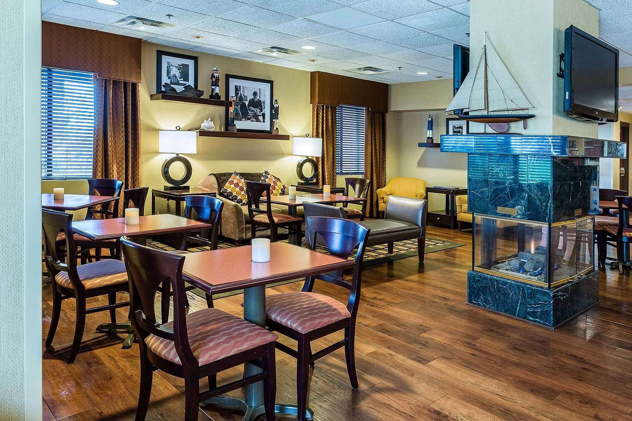 A hotel lobby with wooden flooring, multiple dining tables and chairs, a fireplace, and a television mounted on the wall.