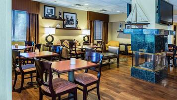 A hotel lobby with wooden flooring, multiple dining tables and chairs, a fireplace, and a television mounted on the wall.