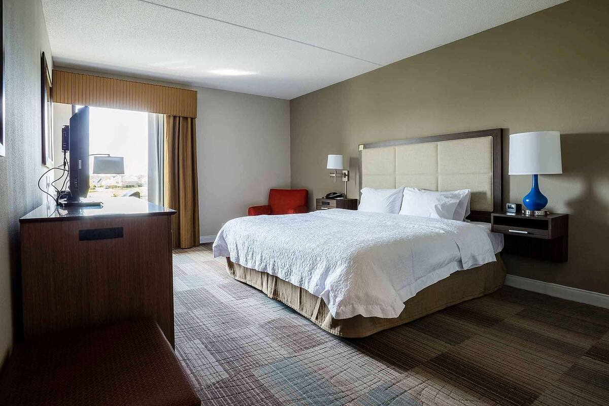 Ein Hotelzimmer mit einem großen Bett, einem Schreibtisch mit Computer, einem Sessel und zwei Nachttischlampen.