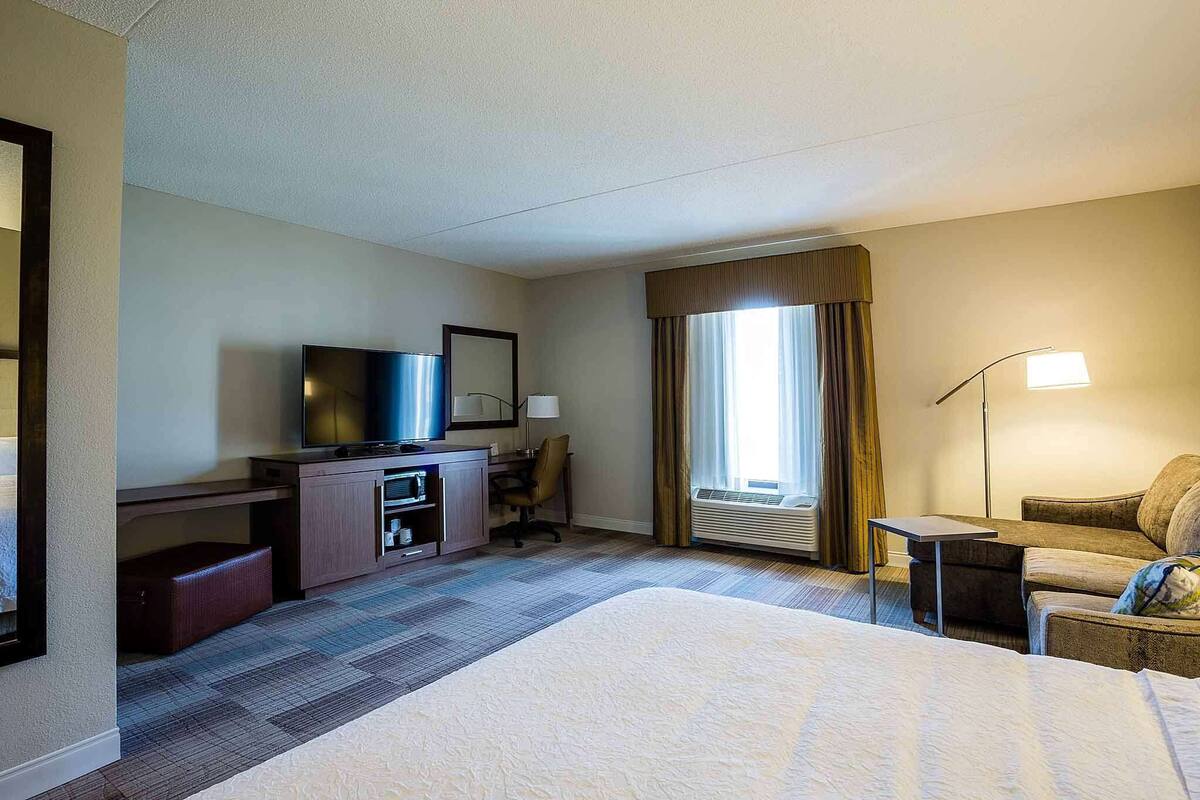 Ein Hotelzimmer mit einem Bett, einem Schreibtisch mit Fernseher, einem Sessel, einer Lampe und einem Fenster mit Vorhängen.