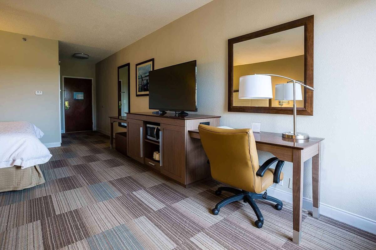Ein Hotelzimmer mit einem Bett, einem Schreibtisch mit Computer, einem Sessel, einem Fernseher und einem Spiegel.