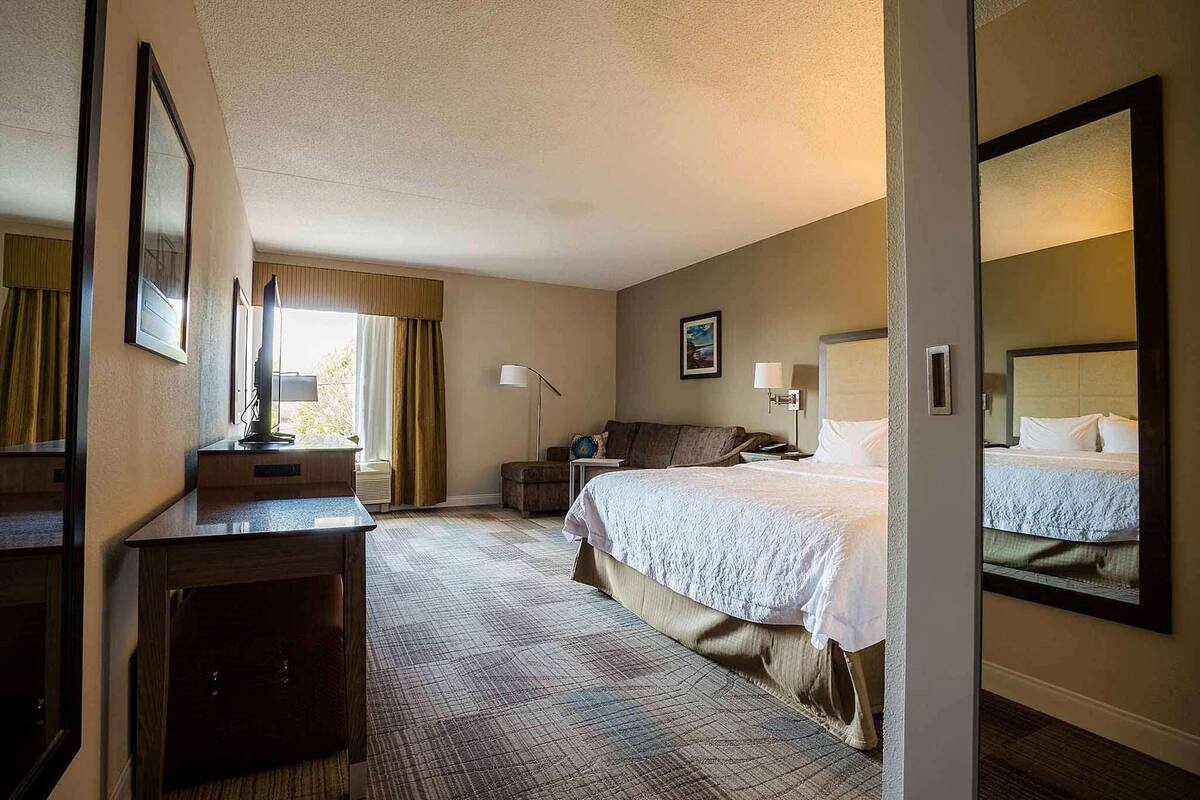 Ein Hotelzimmer mit Bett, Nachttischen, Schreibtisch, Stuhl, Lampe, Fenster mit Vorhängen und Spiegel.