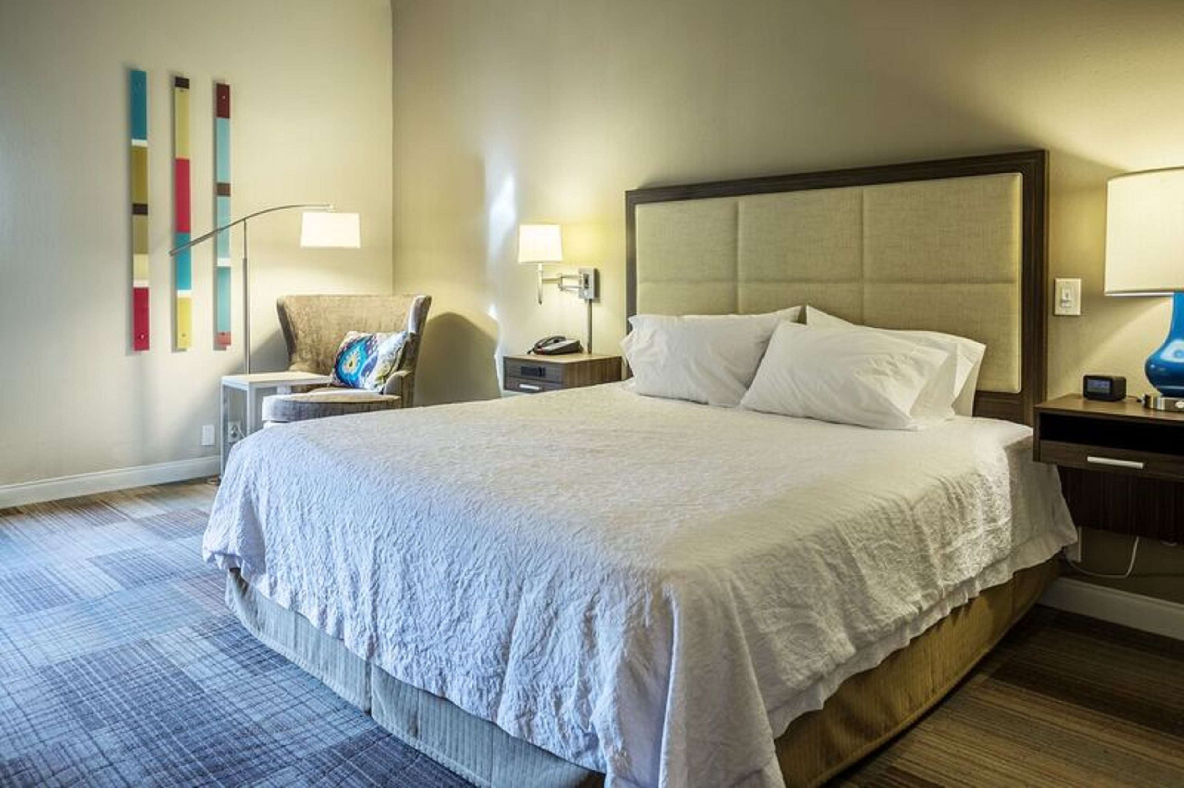 A hotel room with a large bed, a bedside table with a lamp, a chair, and a wall with abstract art.