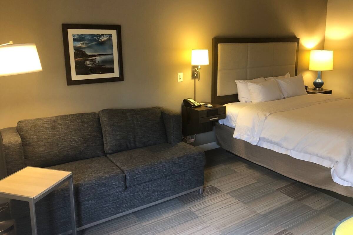 Ein Hotelzimmer mit Bett, Sofa, Nachttisch, Lampe und einem gerahmten Bild an der Wand.