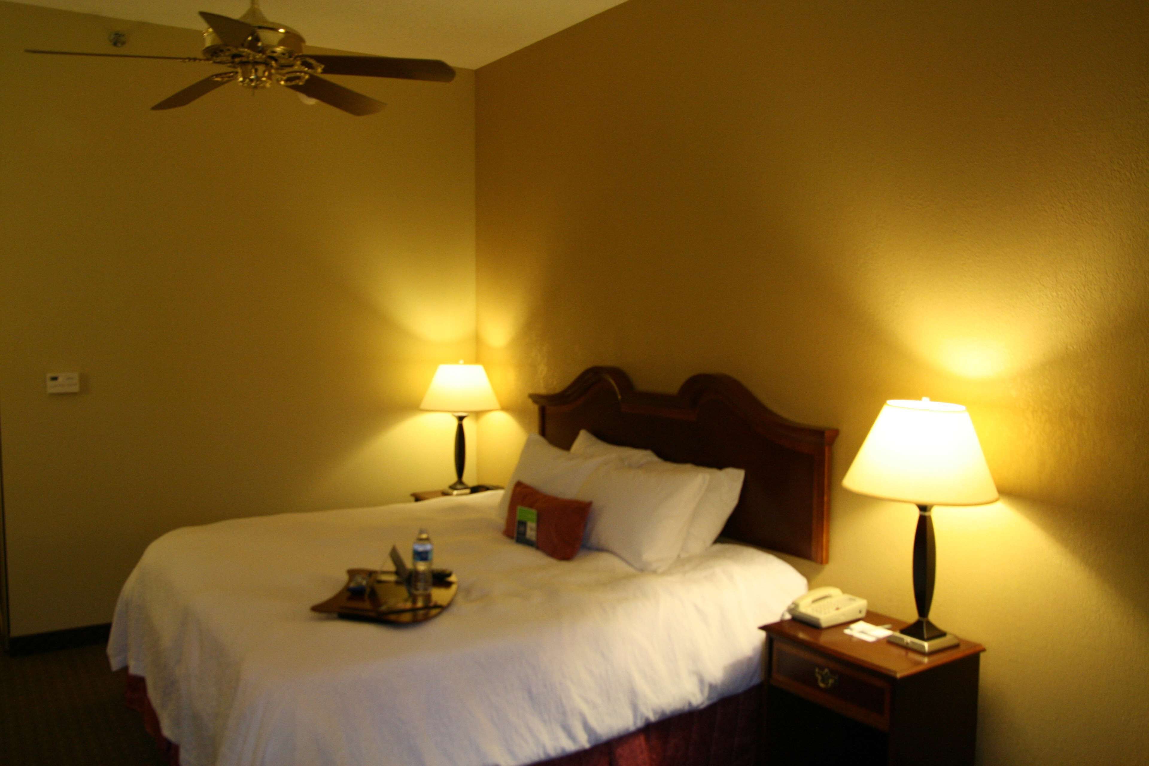 Habitación de hotel con cama, dos lámparas de noche, ventilador de techo y mesita de noche con teléfono y bandeja.