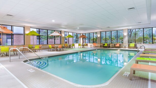 An indoor swimming pool with clear blue water, surrounded by bright yellow and green chairs and tables, and large windows offering a view of the outdoors. - Hampton Inn Boston/Marlborough (Marlborough)
