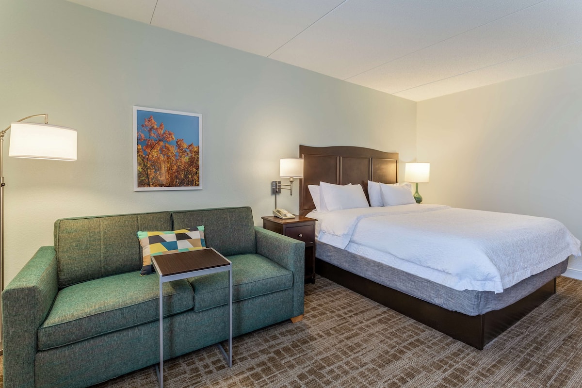 A hotel room with a bed, a green sofa, a small table, a nightstand, and a wall-mounted lamp.