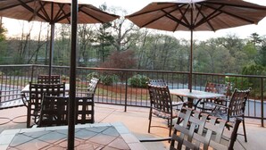 An outdoor patio with tables and chairs, covered by umbrellas, and a view of trees and a road in the background. - Hampton Inn Boston/Peabody (Peabody)