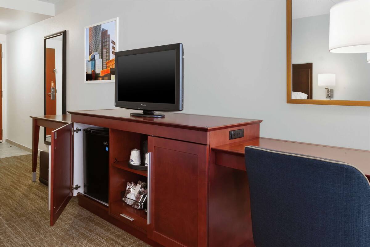 A hotel room with a wooden desk, a television, a minibar, and a mirror.