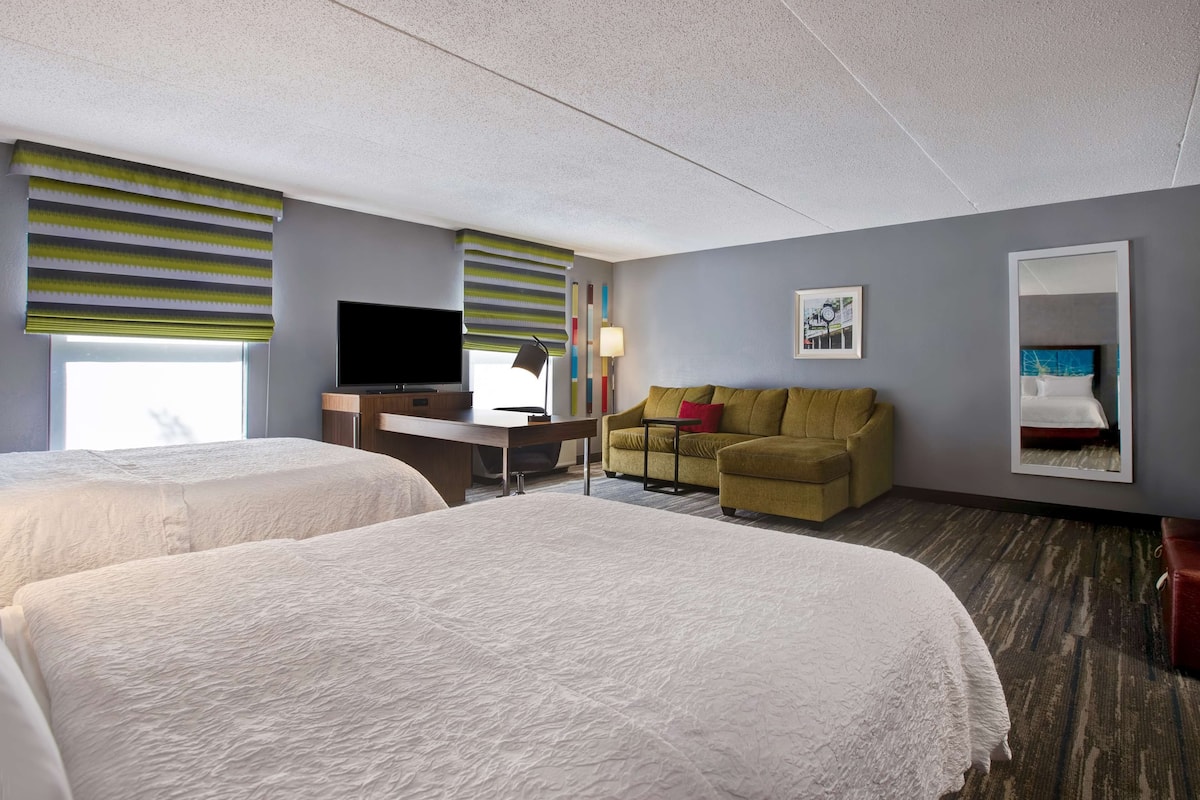 Quarto de hotel com duas camas, uma escrivaninha, televisão, sofá e espelho.