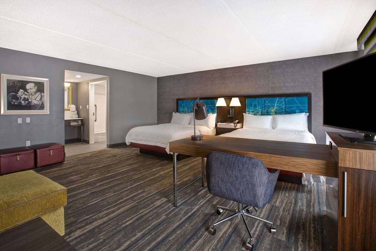 Quarto de hotel com duas camas, uma escrivaninha, uma cadeira, televisão e um quadro na parede.