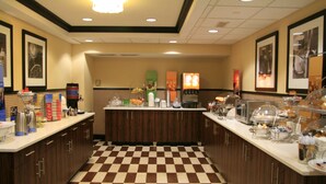 A hotel kitchen with checkered flooring, wooden cabinets, and various food items displayed on the counters. - Hampton Inn Olive Branch (Olive Branch)