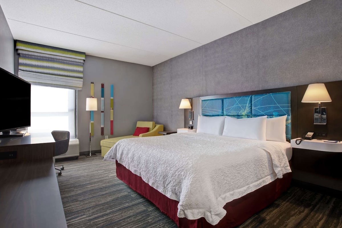 Quarto de hotel com uma cama grande, uma escrivaninha com televisão, uma cadeira, mesa de cabeceira com abajur e janela com persianas.