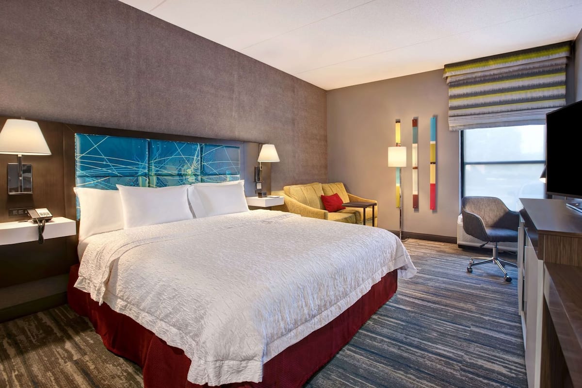 Quarto de hotel com uma cama grande, uma escrivaninha, uma cadeira, um abajur, um telefone e uma janela com vista.