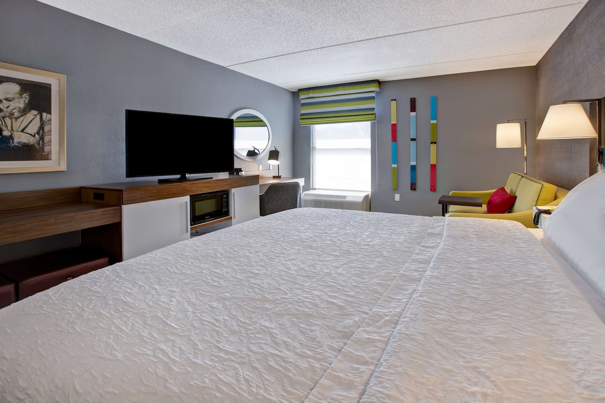 Quarto de hotel com uma cama grande, televisão de tela plana, escrivaninha com computador, cadeira, sofá, janela com persianas e parede com decoração colorida.