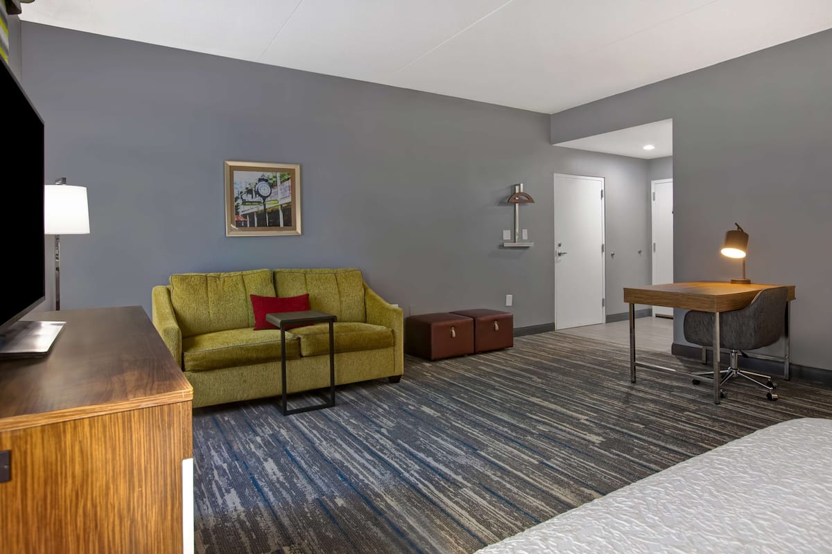 Quarto de hotel com sofá verde, escrivaninha de madeira, luminária, mesa pequena e uma cama.