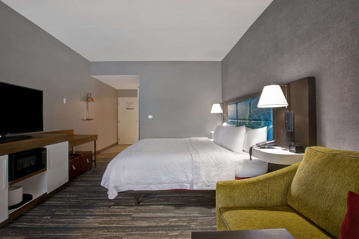 Quarto de hotel com cama, televisão, escrivaninha com micro-ondas, luminária e sofá.