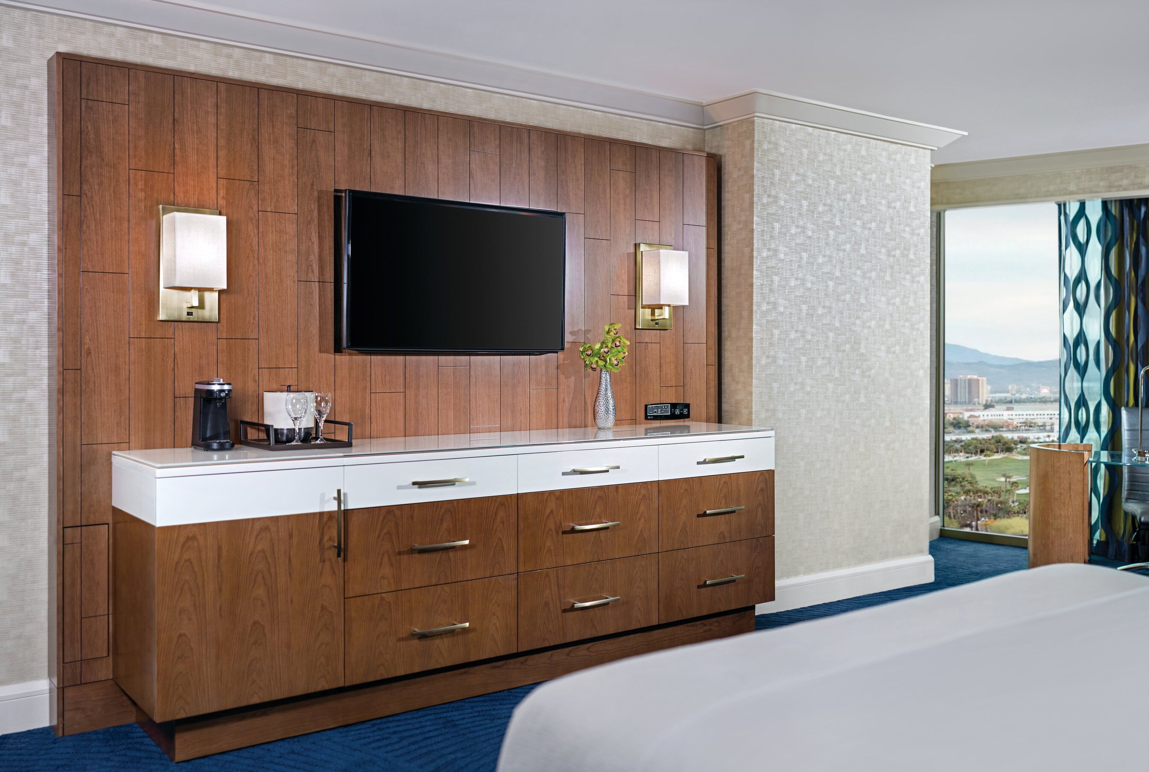 Une chambre d’hôtel dotée d’un grand téléviseur à écran plat fixé sur un mur en panneaux de bois, d’un minibar et offrant une vue sur la ville grâce à de grandes fenêtres.