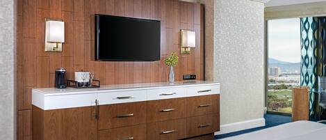 A hotel room with a large flat-screen TV mounted on a wooden panel wall, a minibar, and a view of the city through large windows.