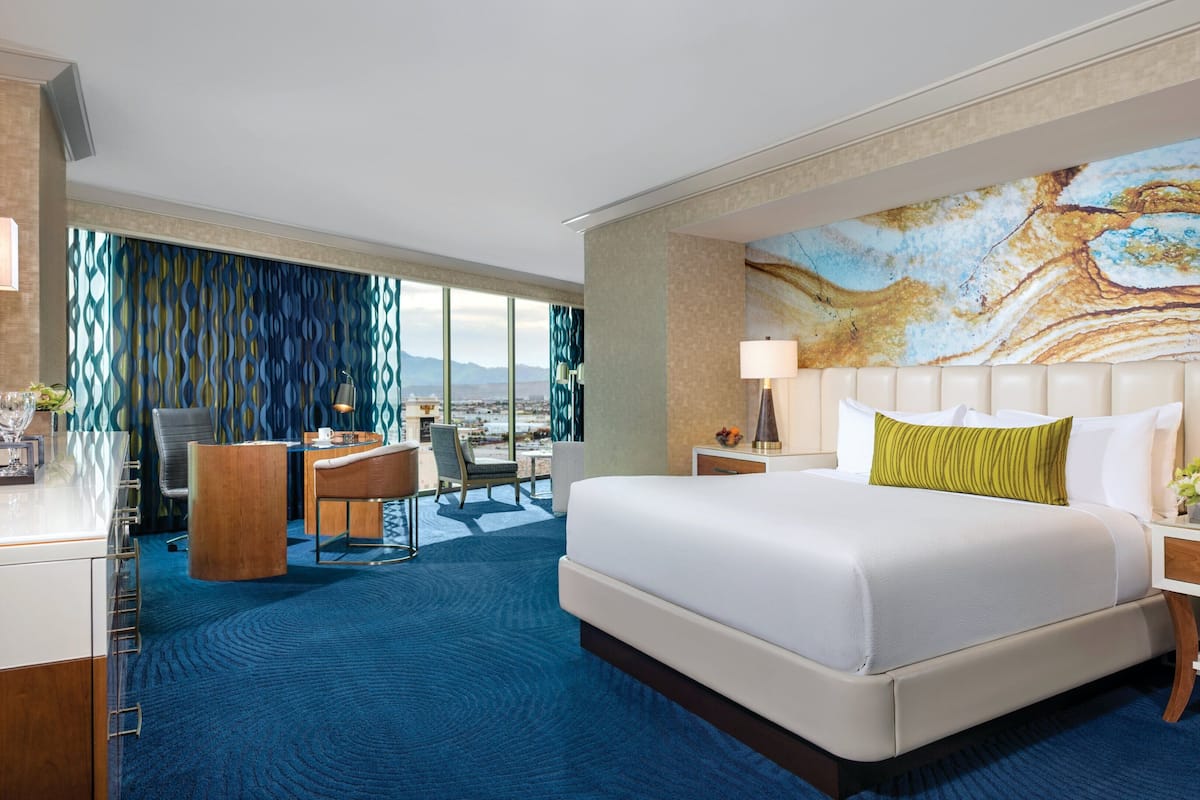 Habitación de hotel con una cama grande, un escritorio con silla, vista a la ciudad y un cuadro abstracto de grandes dimensiones en la pared.