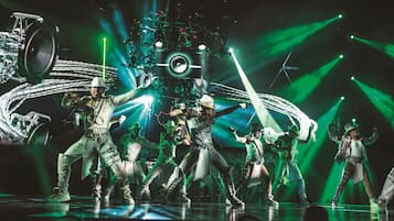 A group of dancers performing on a stage with a large, mechanical backdrop and green lighting.