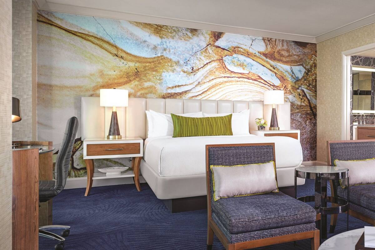 Habitación de hotel con una cama grande, dos sillones, una mesita, un televisor montado en un panel de madera y un baño visible a través de una puerta abierta.