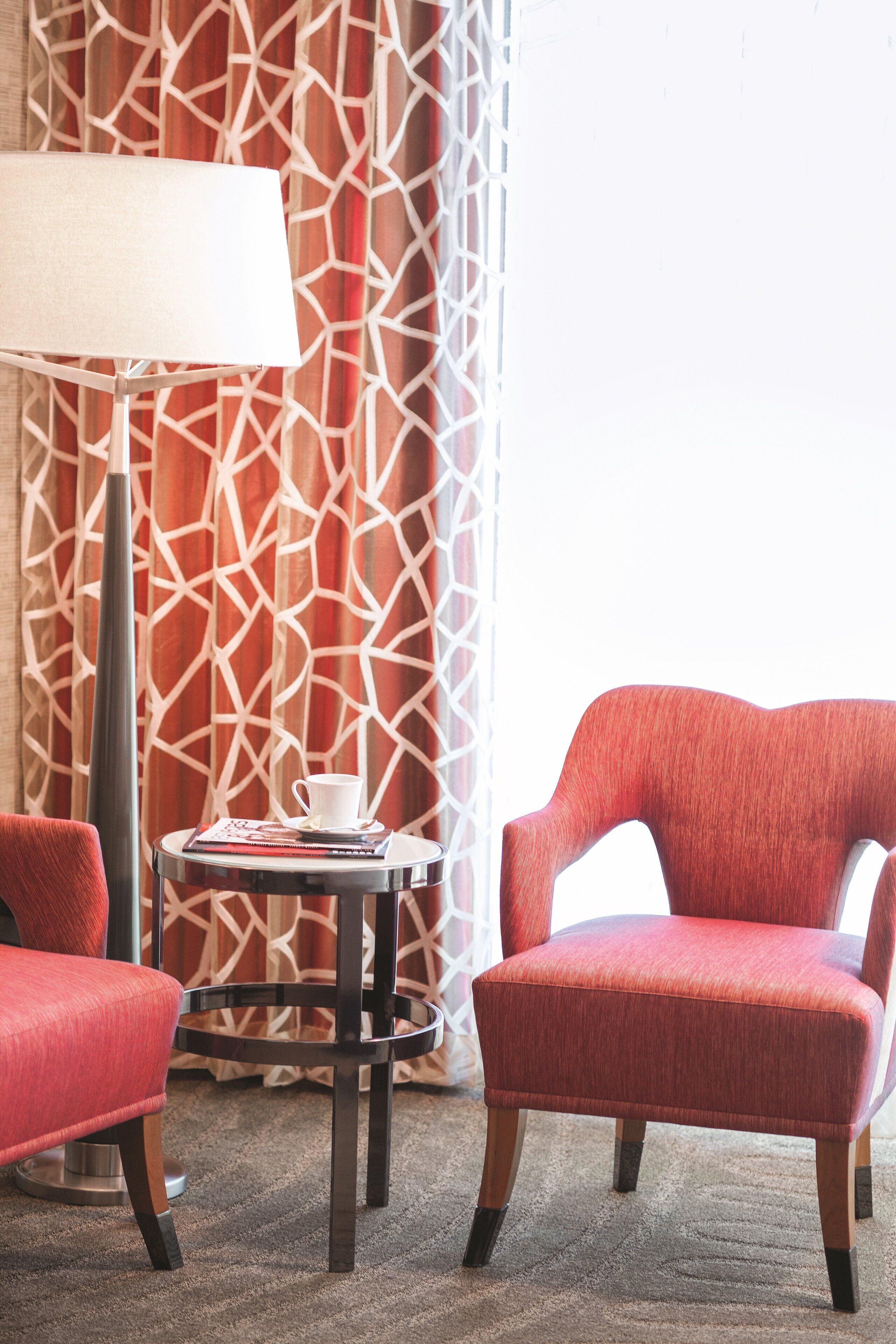 A room with a red chair, a small table with a lamp, and a patterned curtain.
