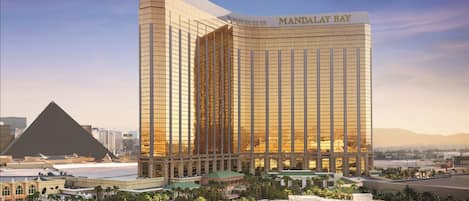 A modern hotel with a prominent glass facade, named "MANDALAY BAY," featuring a large pool area surrounded by palm trees and a desert landscape in the background.