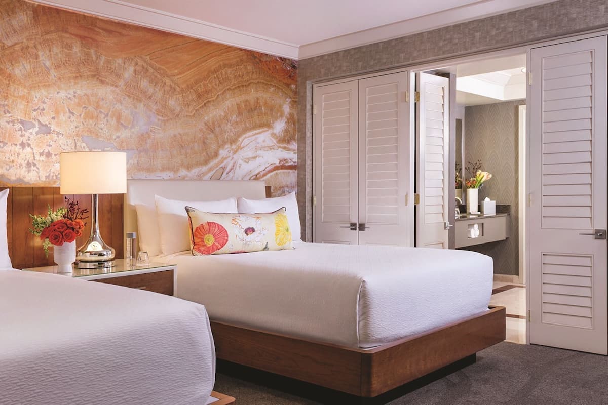 Habitación de hotel con dos camas, una mesita de noche con lámpara, un arreglo floral y un armario con puertas correderas.