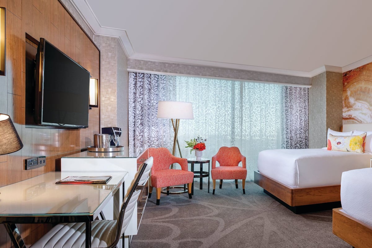 Habitación de hotel con televisión de pantalla plana, escritorio con lámpara, dos sillas rojas, una mesita con un jarrón de flores, un ventanal con cortinas estampadas y una cama con ropa blanca y almohadas decorativas.