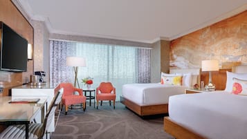 A hotel room with a flat-screen TV, a desk with a lamp, two red chairs, a small table with a vase of flowers, a large window with patterned curtains, and a bed with white bedding and decorative pillows.
