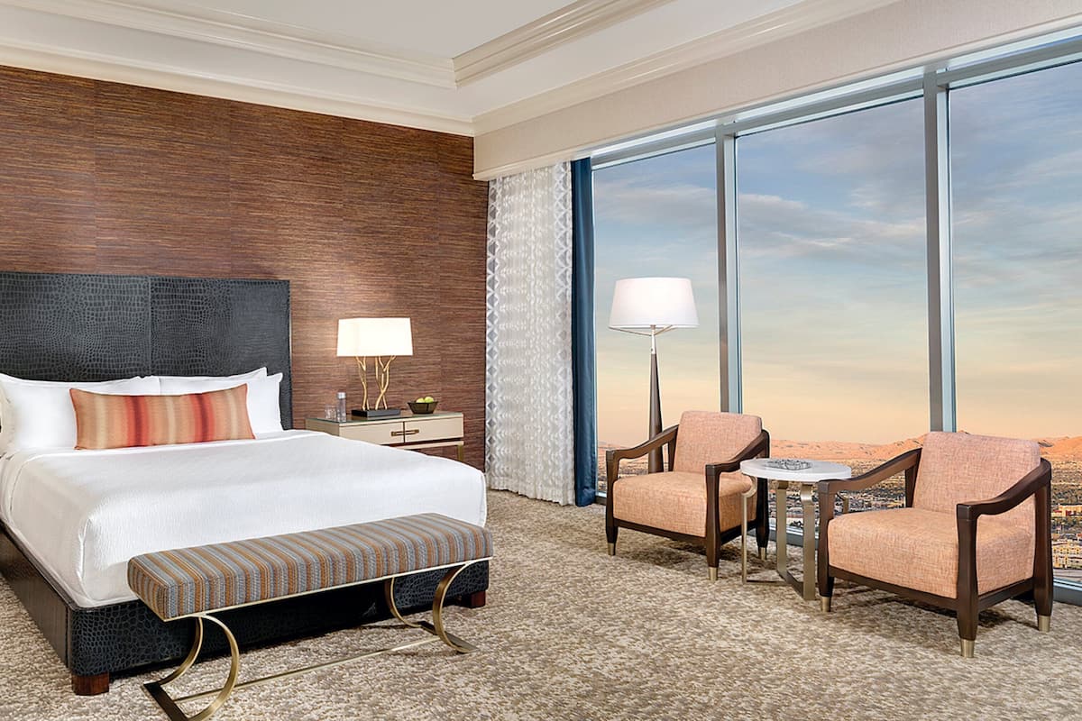Habitación de hotel con una cama grande, dos mesitas de noche con lámparas, un banco, dos sillones y un amplio ventanal con vista a la ciudad.