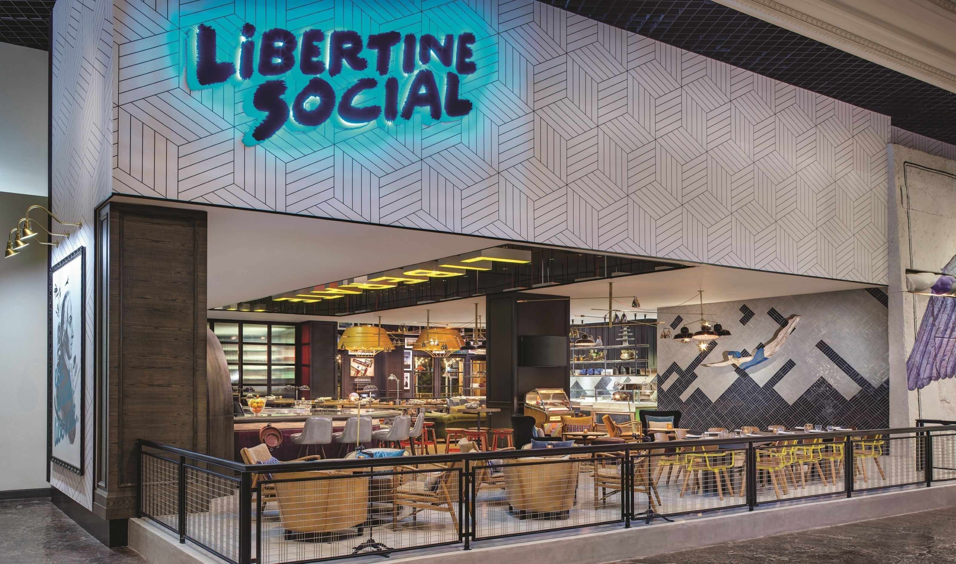 Un interno moderno con parete a motivi a spina di pesce, zona salotto visibile con tavoli e sedie, e insegna luminosa con scritto "Libertine SOCIAL".