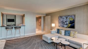 A modern living room with a sofa, ottoman, and coffee table, a kitchen area with bar stools, and a large abstract painting on the wall.