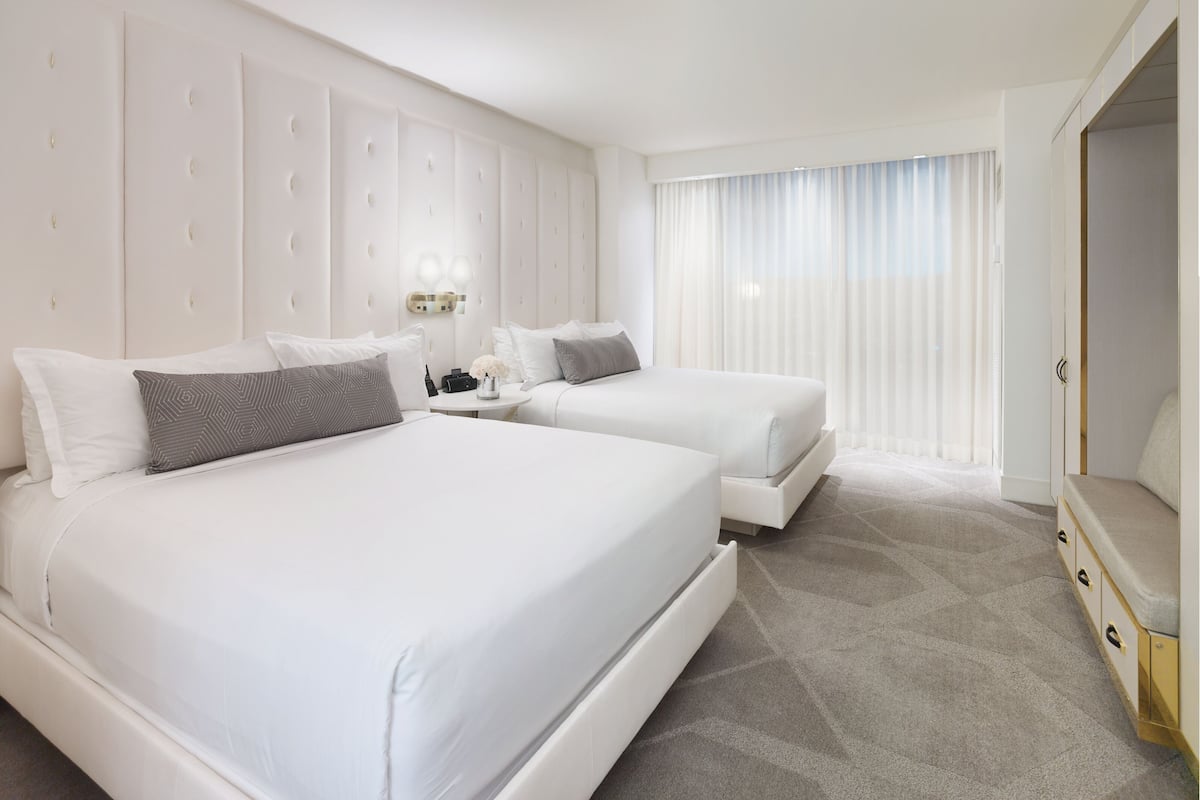 Habitación de hotel con dos camas, un cabecero grande y acolchado, una mesita de noche con lámpara y un ventanal con cortinas translúcidas.
