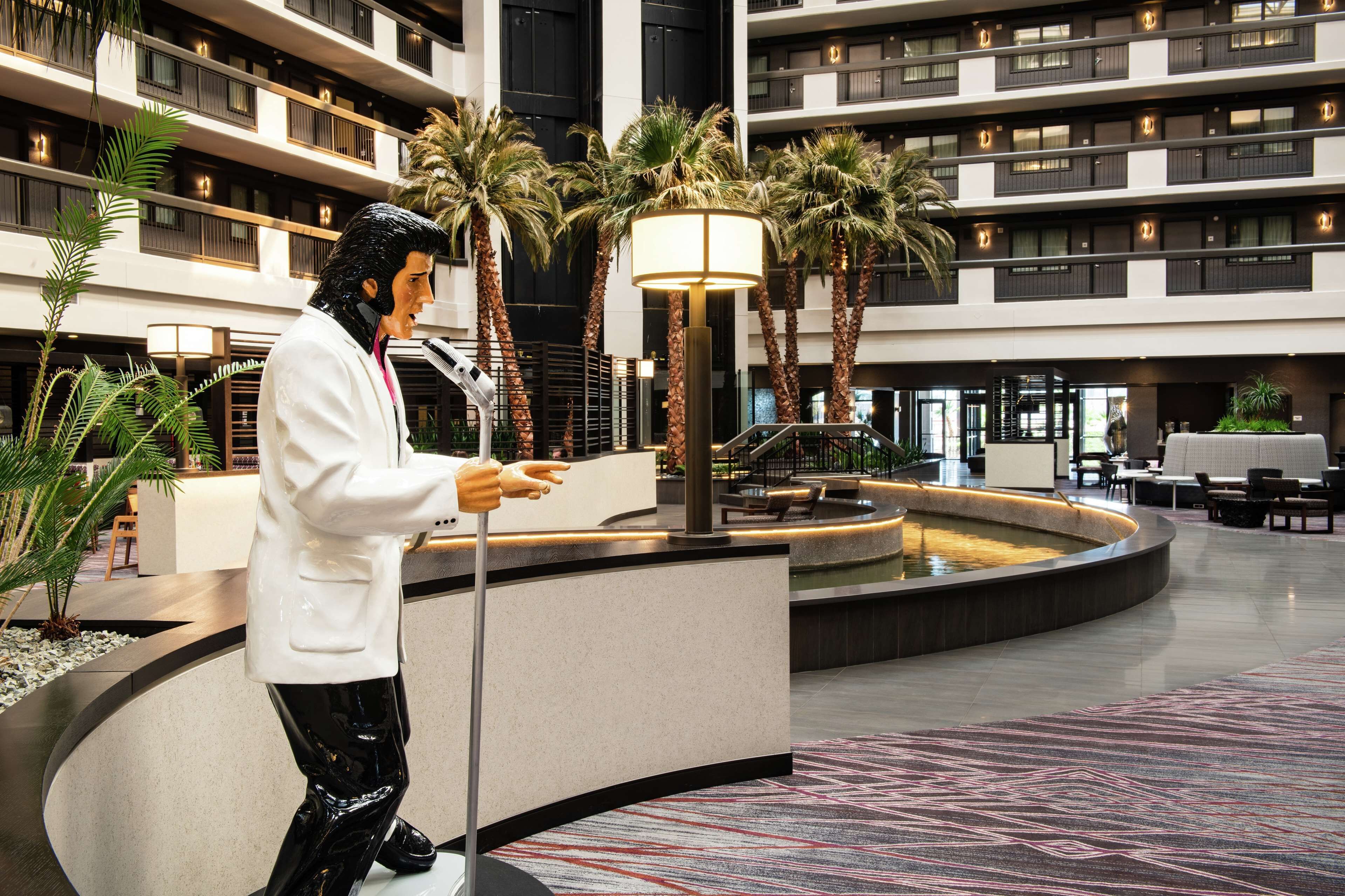 Een moderne hotelentree met een centrale fontein, palmbomen en een standbeeld van een man in pak.