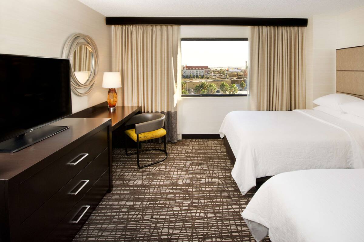 Een hotelkamer met twee bedden, een televisie, een bureau met lades, een stoel en uitzicht op de stad.