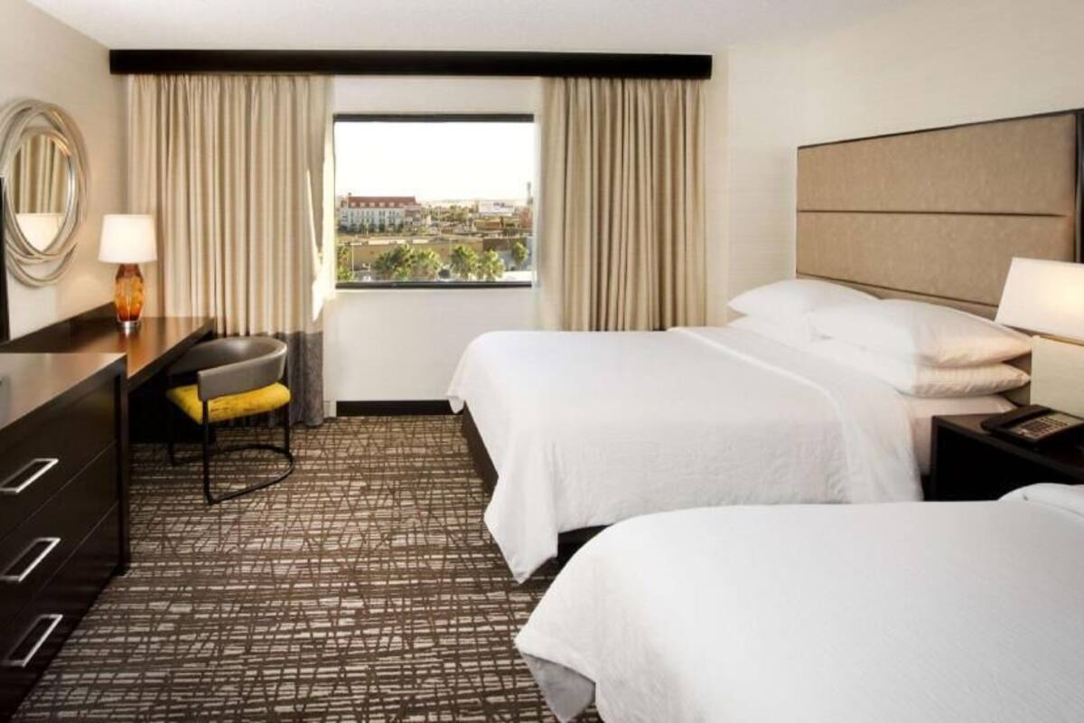 Een hotelkamer met twee bedden, een bureau met een televisie, een stoel, een lamp en een raam met gordijnen.